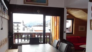 Un coin repas avec une table et des chaises, un canapé rouge et une vue sur les bâtiments et les montagnes, visible à travers une grande fenêtre.