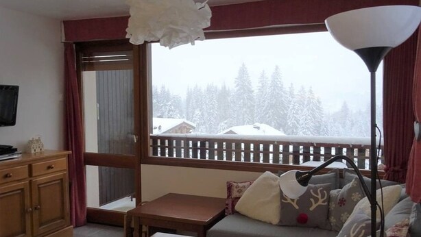 A cozy living room with a sofa, a wooden table, a lamp, and a view of a snowy landscape through a large window.