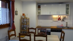 A compact kitchen with white cabinets, a wooden dining table with chairs, and a window with lace curtains.