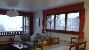 A cozy living room with a sofa, dining table, and chairs. There is a window with curtains and a view of trees.