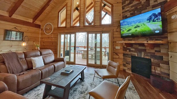 L’intérieur chaleureux d’un chalet avec un plafond en bois, un grand téléviseur fixé à un mur en pierre, un canapé en cuir et une porte vitrée donnant sur une terrasse extérieure.