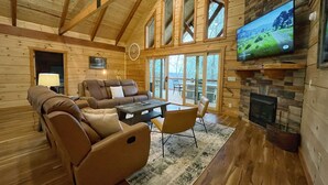 L’intérieur d’un chalet confortable, avec une cheminée, un grand téléviseur fixé au mur et un coin salon accueillant, comprenant un canapé et des fauteuils.