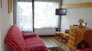 Una habitación con sofá rojo, cómoda de madera, una mesita, televisor montado en la pared y ventana con cortinas.