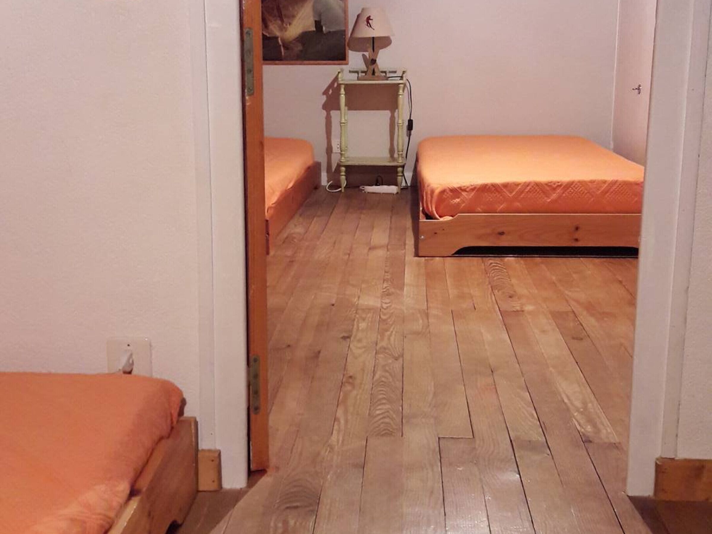 Un couloir étroit avec deux lits en bois, chacun recouvert d’une couverture orange. On aperçoit une petite table avec une lampe au fond.
