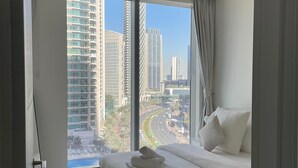Une chambre moderne avec un grand lit, une vue sur le paysage urbain grâce à de grandes fenêtres du sol au plafond, et un luminaire au plafond.