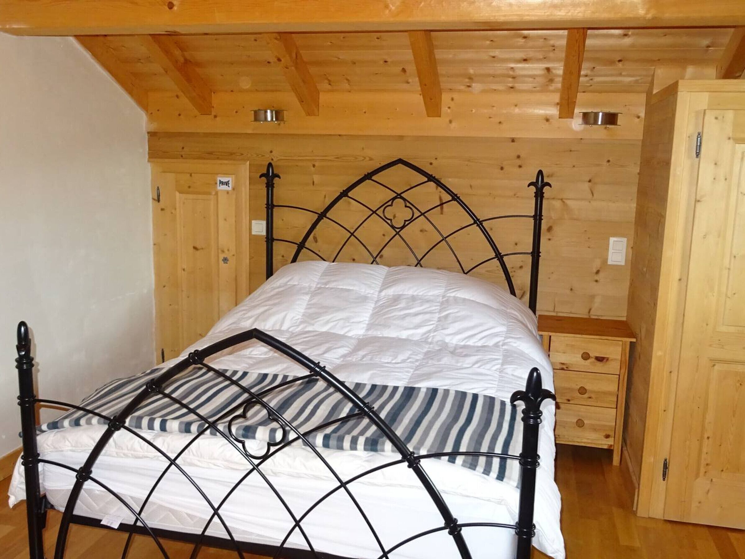 Uma cama de solteiro com estrutura de metal e colchão acolchoado num quarto com paredes de madeira e teto inclinado.