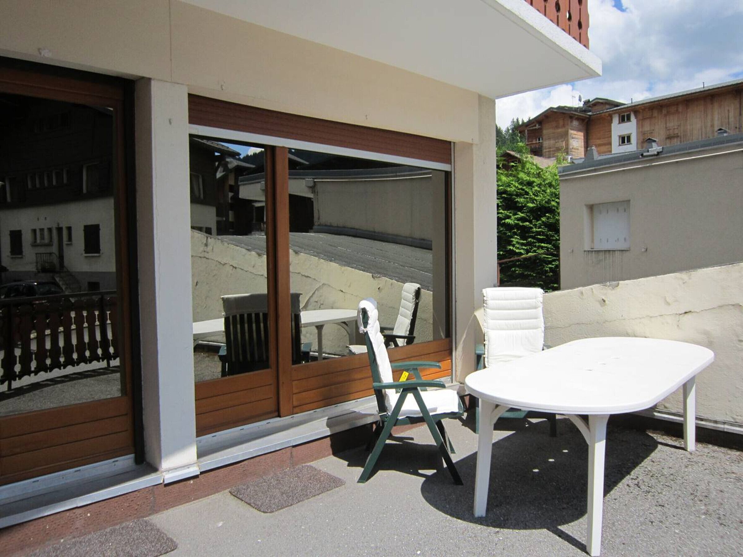 A patio area with a white table and two chairs, a sliding glass door, and a view of a street and buildings.
