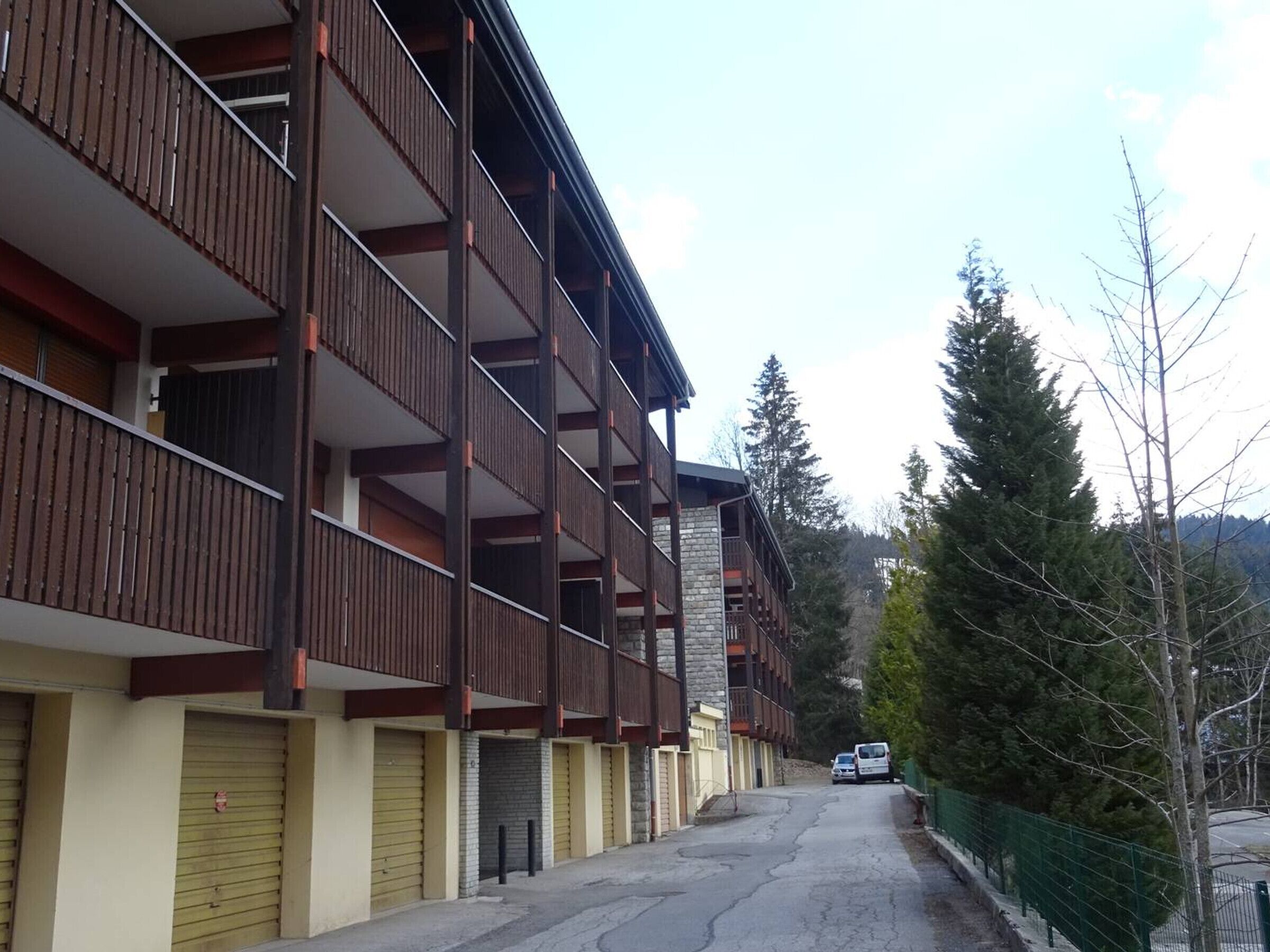 A multi-story building with balconies, a parking area, and a forested backdrop.