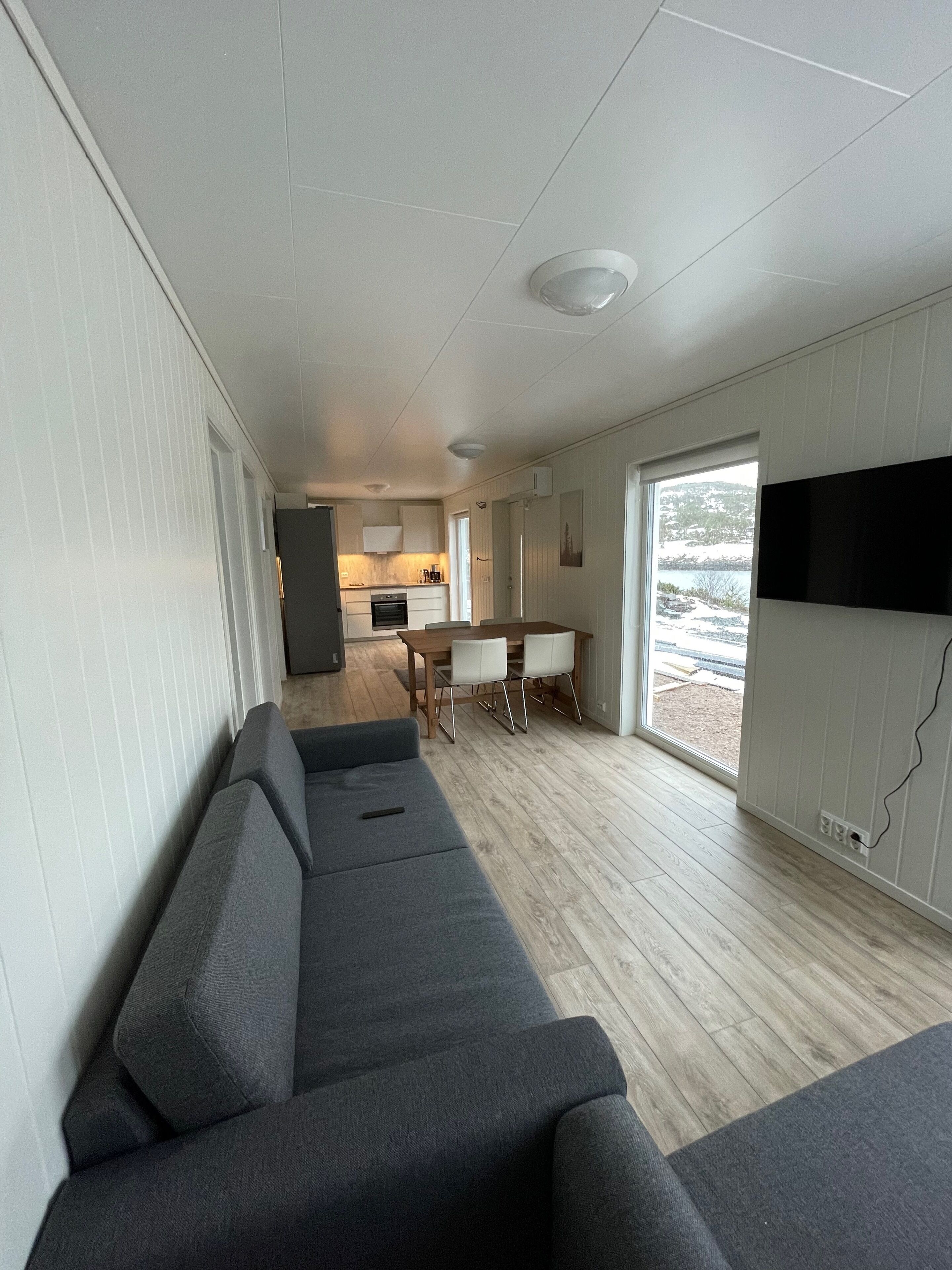 Ett modernt vardagsrum med en grå soffa, ett matbord med stolar, en köksdel och ett stort fönster med utsikt över omgivningen.