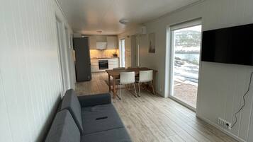 Ett modernt vardagsrum med en grå soffa, ett matbord med stolar, en köksdel och ett stort fönster med utsikt över omgivningen.