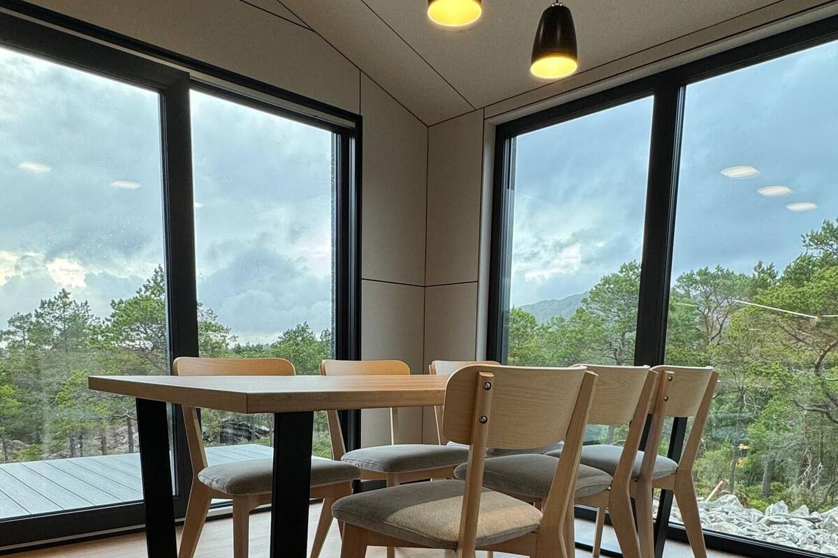 A modern dining area with a wooden table and chairs, large windows with a view of trees and mountains, and pendant lighting.