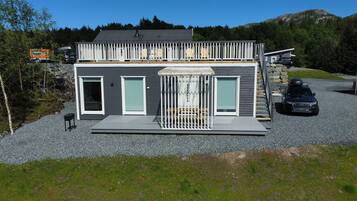 A modern two-story cabin with a deck, a covered outdoor seating area, and a gravel driveway.
