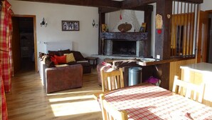 Un salon chaleureux avec une cheminée, des poutres apparentes, un canapé, une table recouverte d’une nappe à carreaux et un radiateur.