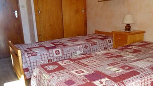 A bedroom with two wooden beds, each covered with a patterned quilt. There is a wooden nightstand with a lamp and a wooden wardrobe with a mirror. - Cozy 2-Room Apartment for 5, South-Facing Balcony, Near Shops and Slopes, Garage Included (Les Gets)