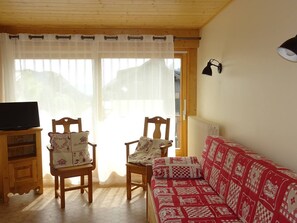Ein gemütliches Wohnzimmer mit zwei Holzstühlen, einem rot-weißen Sofabezug und einem Fernseher auf einer Holzkommode. Ein Fenster mit transparenten Vorhängen.