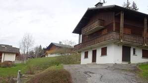 A two-story wooden house with a balcony, a driveway leading up to it, and a grassy hill in the background. - Les Gets Duplex 3-Room - 6 Pax, Comfortable, Garage (Les Gets)