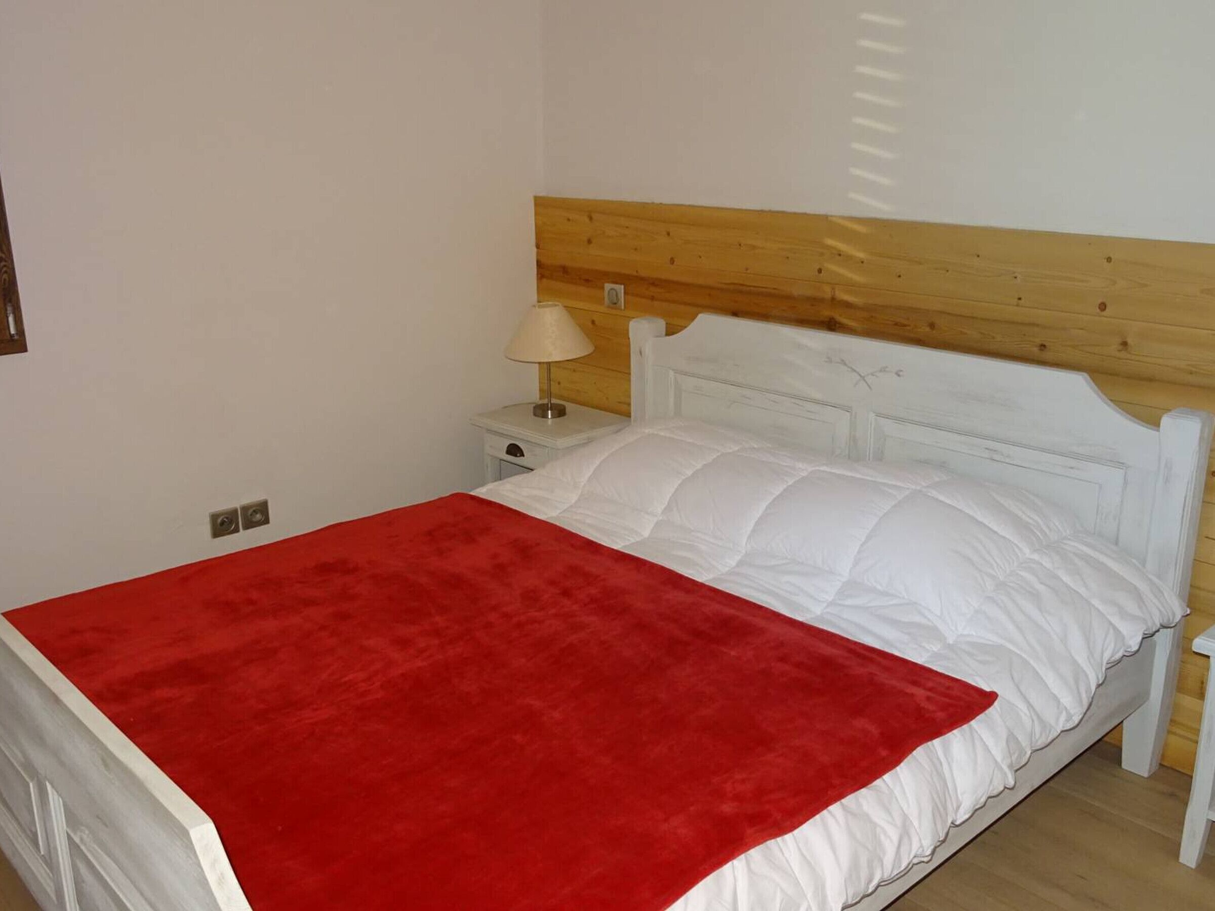 Una cama con una manta roja, almohadas blancas y un cabecero de madera.