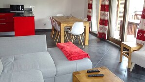 Un espace de vie moderne avec un canapé gris, une table à manger en bois avec des chaises, un ensemble de cuisine rouge et une grande fenêtre ornée de rideaux rouges et blancs.