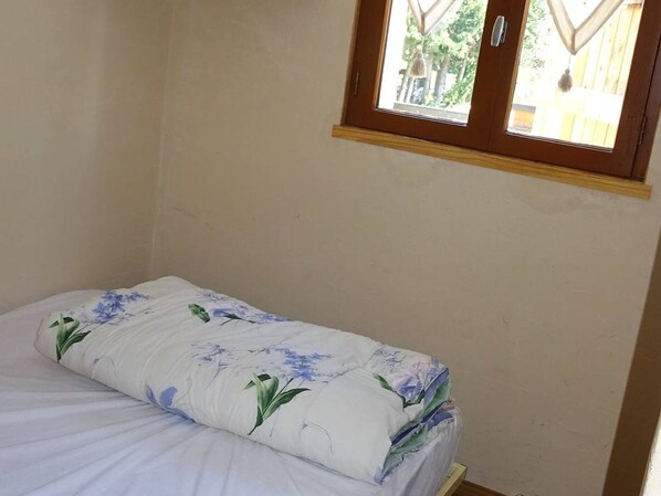 Una habitación pequeña con una cama individual y una colcha de estampado floral, un marco de ventana de madera y una pared lisa.