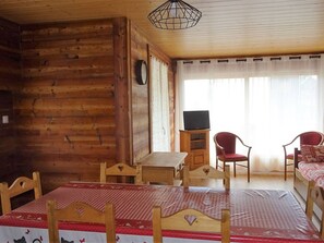 A cozy cabin room with a wooden interior, a bed with a checkered bedspread, two chairs, a small table, a television on a stand, and a window with sheer curtains.