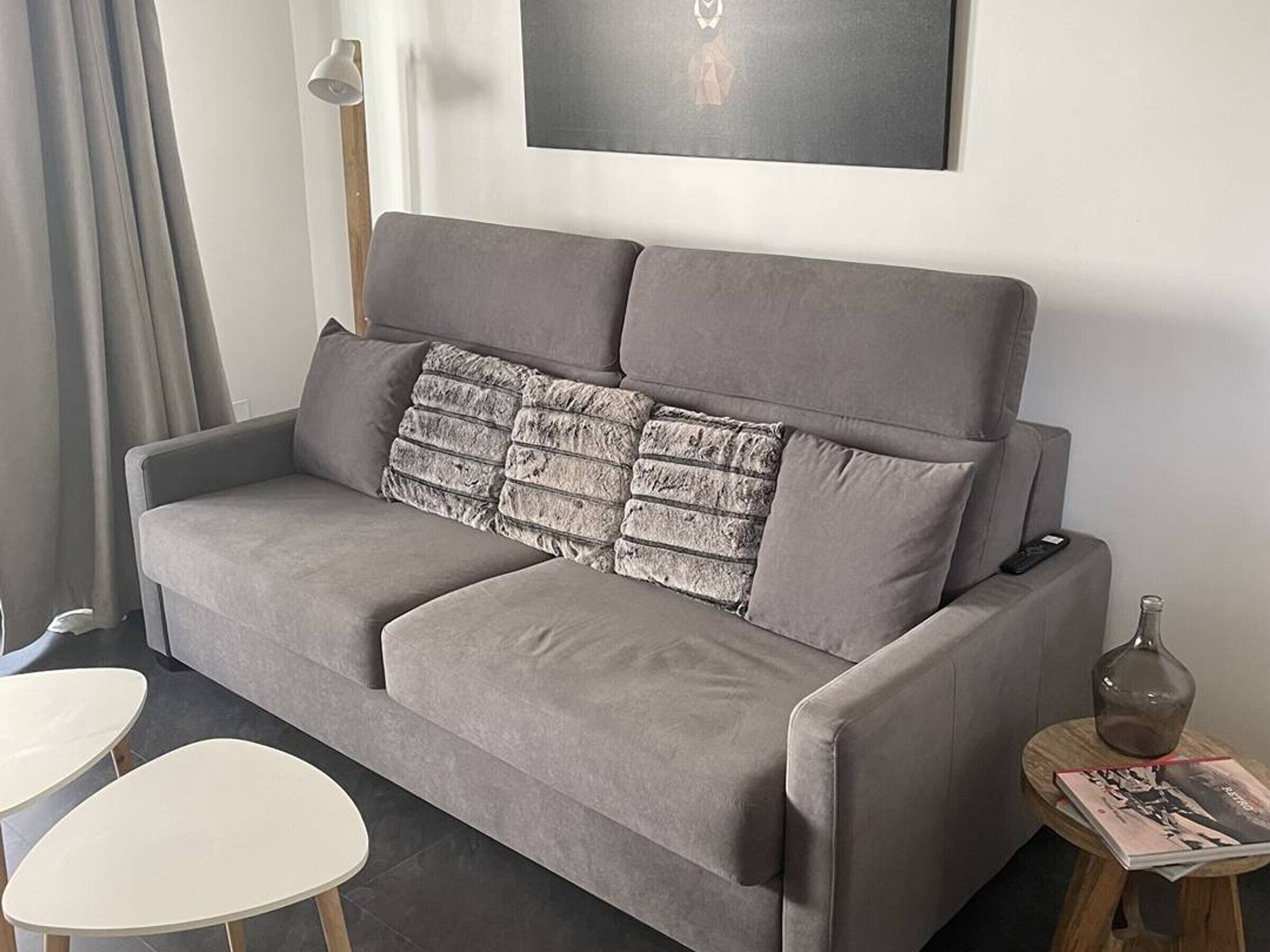 A grey sofa with a textured throw pillow, a small round table with a bottle and magazine, and a white table.