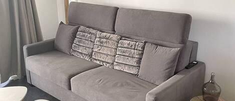 A grey sofa with a textured throw pillow, a small round table with a bottle and magazine, and a white table.