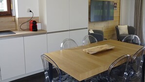 Una cocina moderna con armarios blancos, encimera de madera y una cafetera roja. Hay una zona de comedor con una mesa de madera y sillas transparentes.