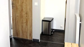 Un pasillo estrecho con una puerta de madera a la izquierda, un bote de basura pequeño en el centro y una pared blanca.