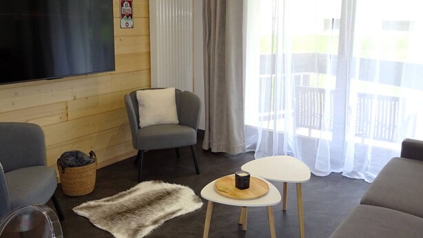 Una sala de estar acogedora con una televisión de pantalla plana, sillones grises, una mesita redonda con una vela y una alfombra de piel en el suelo.
