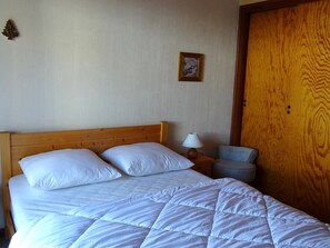 A bedroom with a wooden headboard, a bed with white bedding, a nightstand with a lamp, a small chair, and a wooden door.