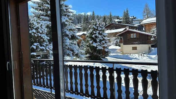 A view from a window with a wooden railing, snow-covered trees, and a building with a red roof. - Spacious 6-Room Duplex, 12 Guests, WiFi, Garage, Close to Slopes - Sheets & Towels Included in Winter (Les Gets)