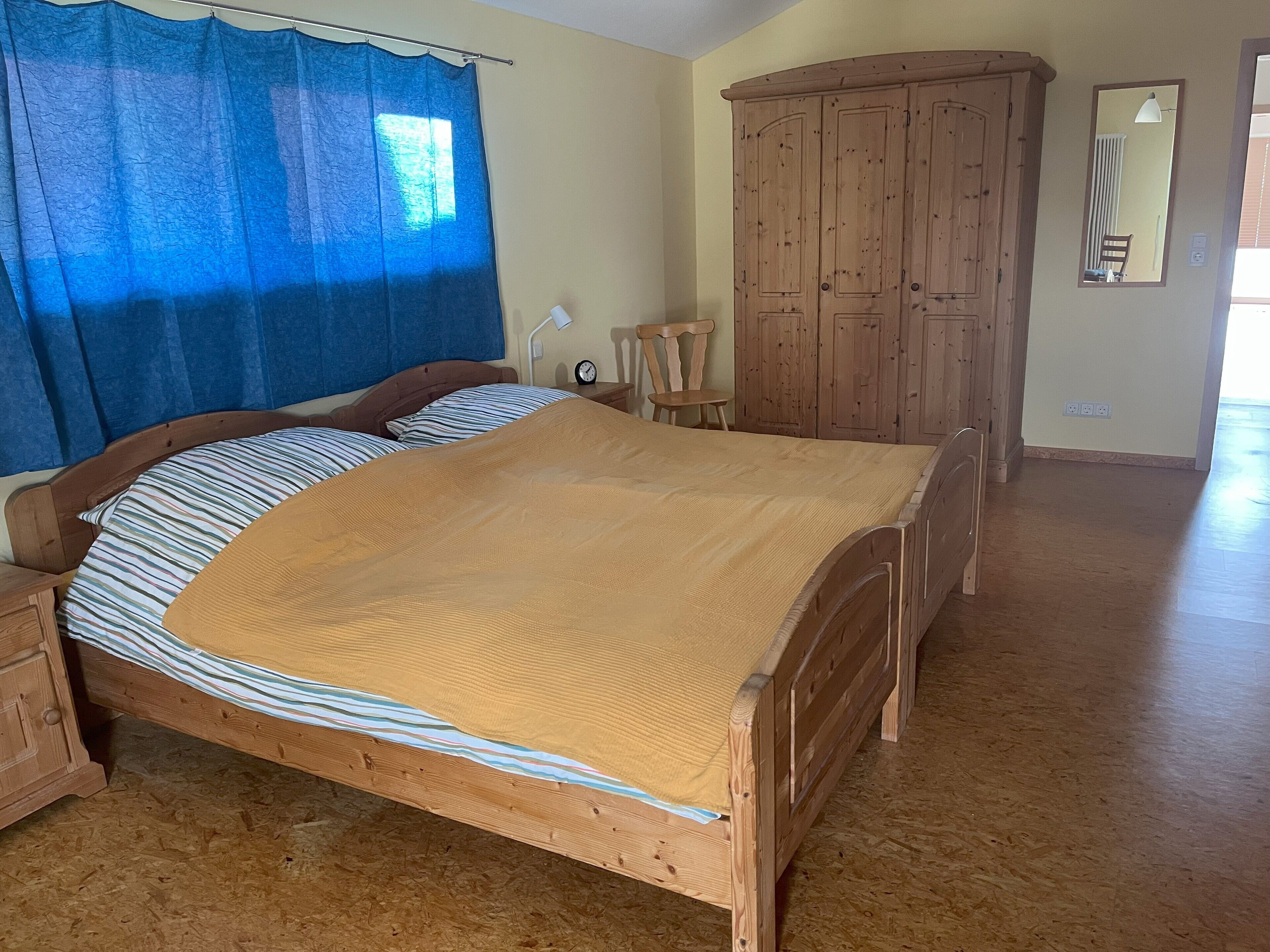 A wooden bed with a striped pillow and a brown blanket, a wooden wardrobe, a wooden chair, and a mirror on the wall.