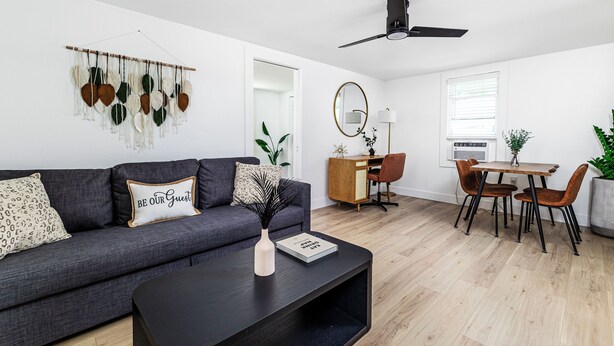Ein modernes Wohnzimmer mit einem dunkelgrauen Sofa, einem schwarzen Couchtisch, einem Essbereich mit einem Holztisch und zwei Stühlen sowie einem Deckenventilator.