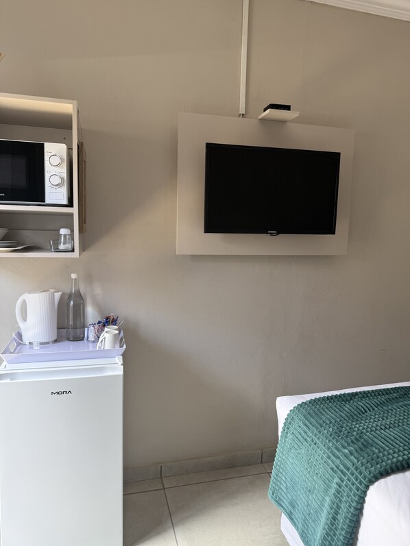 Une chambre avec un lit, un téléviseur fixé au mur, un petit réfrigérateur et une étagère avec une bouilloire et d’autres objets.