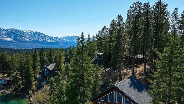 Eine Hütte mit einer Terrasse, umgeben von dichtem Wald und einem See, mit Bergen im Hintergrund.
