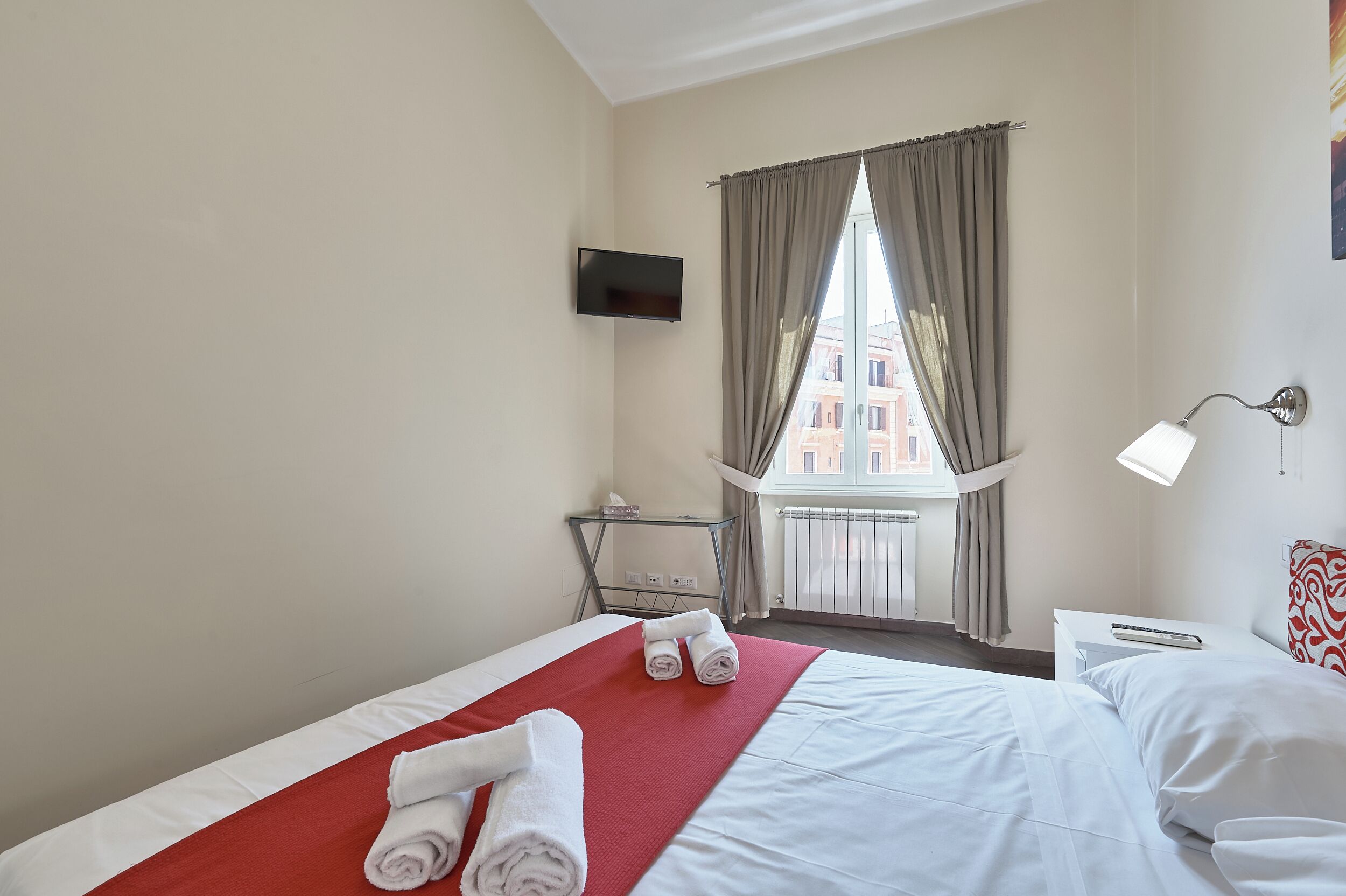 A hotel room with a bed, a TV mounted on the wall, a window with curtains, a bedside table with a lamp, and a view of buildings outside.
