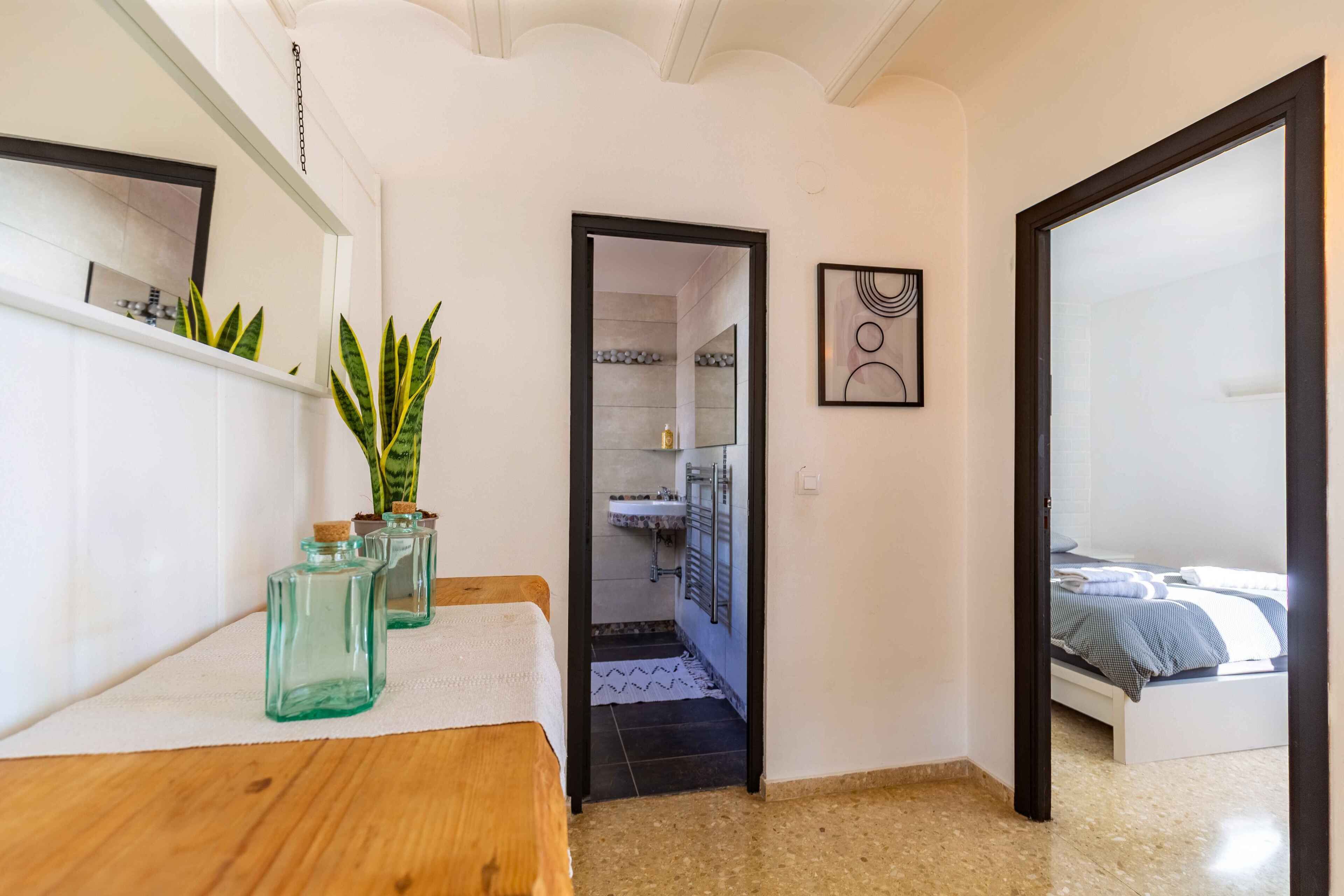 A bedroom with a bed, a wooden bench, and a small table with potted plants. There is a bathroom visible through an open door.