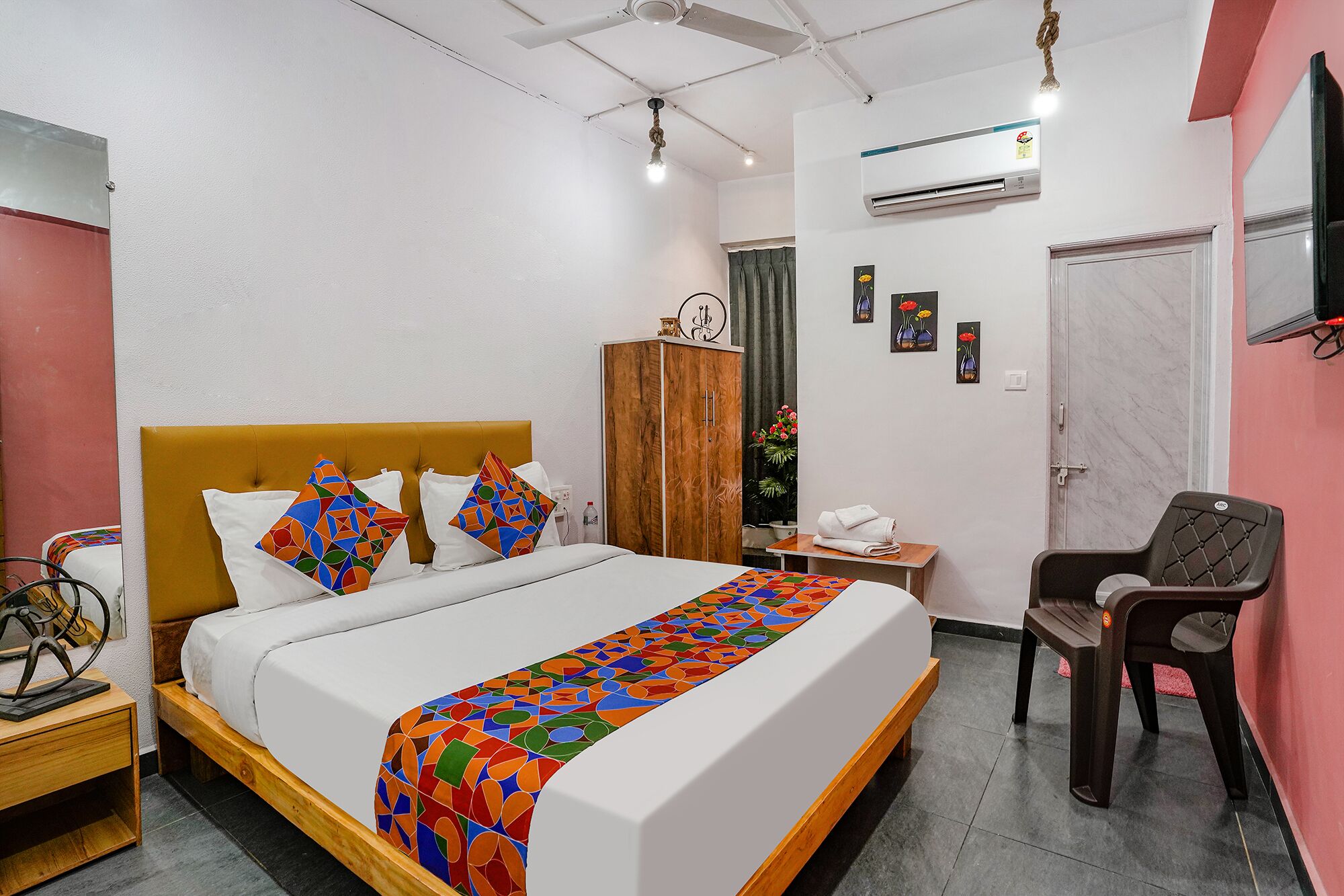 A hotel room with a bed, a wooden nightstand, a desk, a chair, an air conditioning unit, and a wardrobe.
