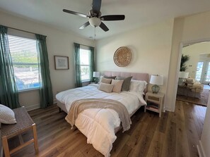 Un dormitorio con cama, ventilador de techo, mesita de noche con lámpara, una silla y ventana con cortinas.
