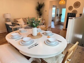 Una mesa de comedor dispuesta con platos, tazones y cubiertos blancos, rodeada por un protector de mesa tejido y un florero con follaje.