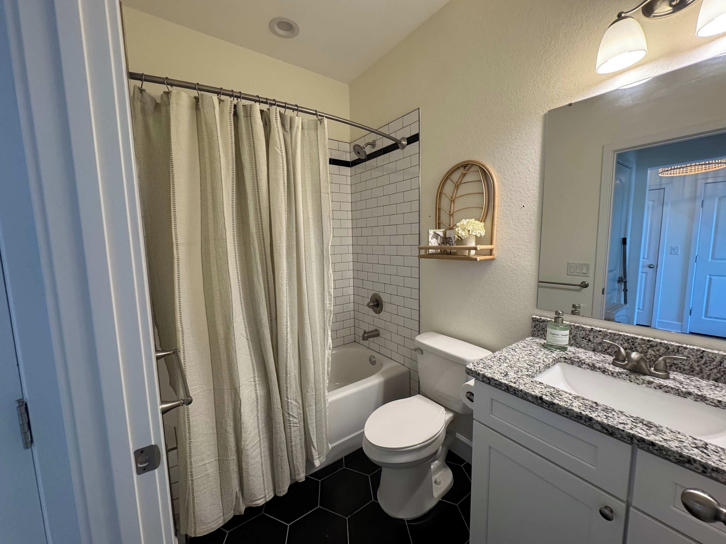 Een badkamer met douchegordijn, bad, toilet en een wastafel met een granieten aanrecht, en een spiegel met een deur op de achtergrond.