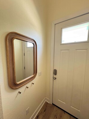 Un couloir avec un miroir encadré en bois, une porte blanche avec une fenêtre et un interrupteur sur le mur.