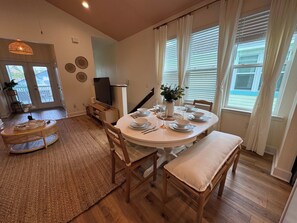 Un coin repas avec une table ronde dressée pour quatre personnes, un téléviseur posé sur un meuble, et une vue sur l’extérieur grâce à de grandes fenêtres.