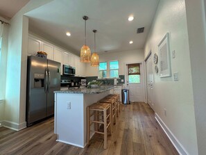A modern kitchen with stainless steel appliances, a central island with stools, pendant lighting, and a view of the outdoors through windows.