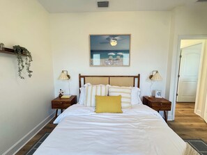 Ein Schlafzimmer mit einem Bett, zwei Nachttischen, einer Wandlampe, einem gerahmten Bild und einer hängenden Pflanze.