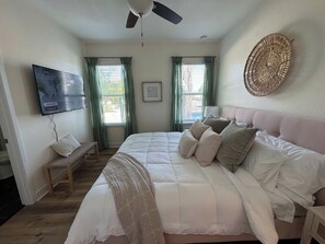A bedroom with a large bed, a ceiling fan, two windows with curtains, a wall-mounted TV, and a woven wall decoration.