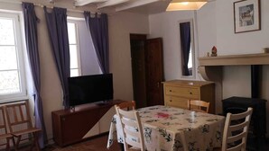 Habitació amb televisió sobre un taulell de fusta, taula amb cadires, llar de foc i finestra amb cortines.