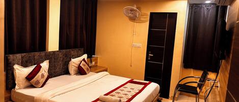 A hotel room with a large bed, dark curtains, a ceiling fan, a wooden nightstand, a TV, and a built-in wardrobe.