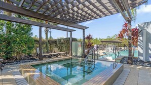Una piscina con una pérgola de madera sobre ella, rodeada de plantas tropicales y un cielo despejado.