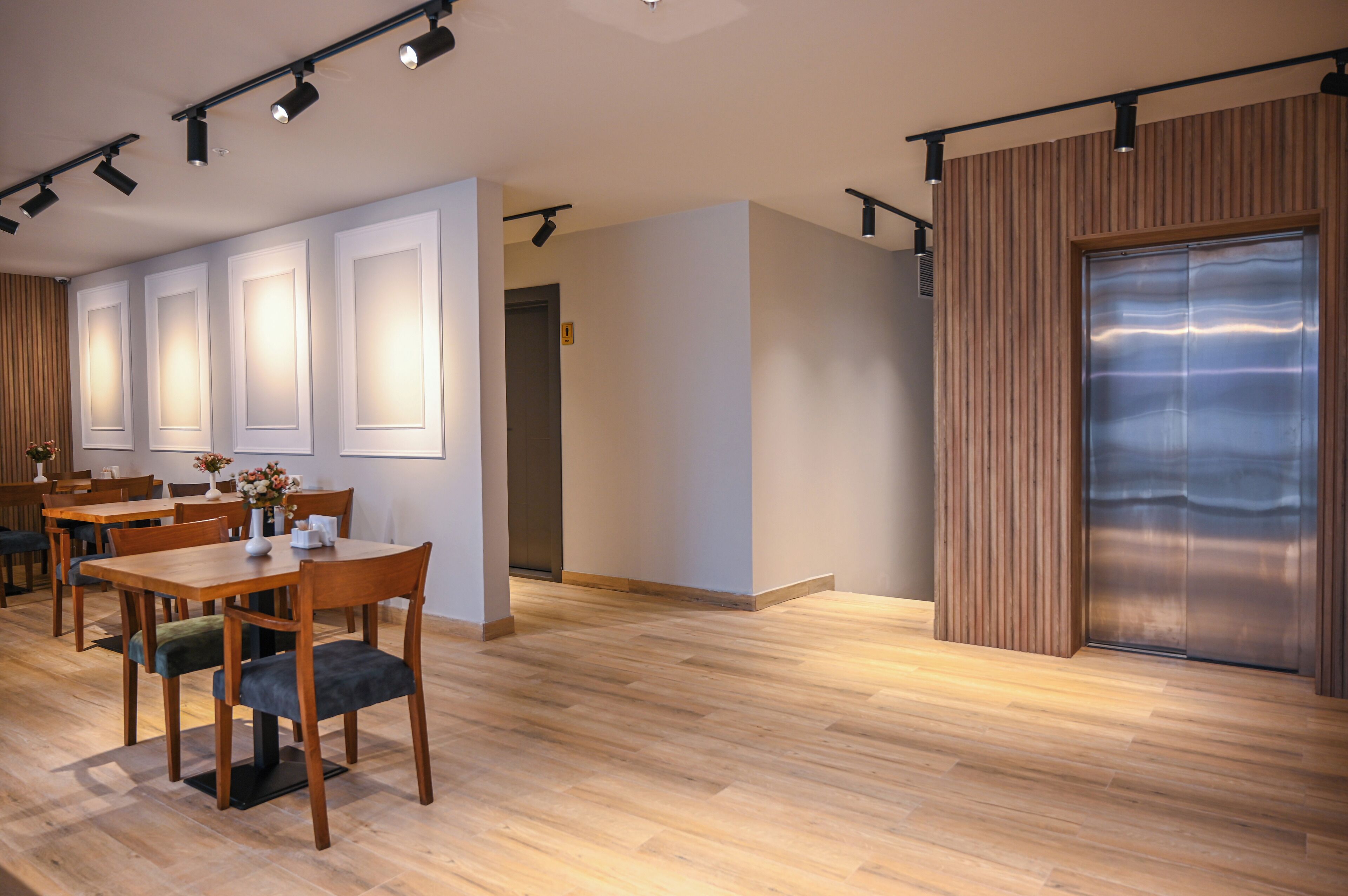 A modern interior with wooden flooring, a dining area with tables and chairs, and a large metallic elevator.